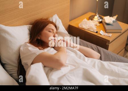 Top view of sick calm redhead young woman wearing soft night clothes sleeping in comfortable bed lying down on soft pillow. Stock Photo