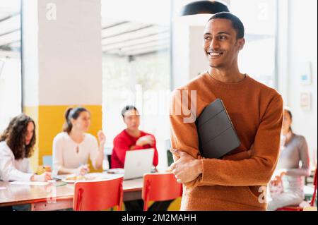 Attractive Moroccan young male in casual clothes standing and looking at his side smiling while holding a tablet. Teamwork sitting at desk on backgrou Stock Photo