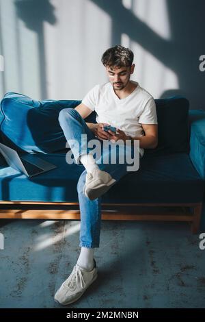 Concentrated young man typing on mobile phone while sitting on a sofa at home near laptop computer. obile phone communication. Education internet Stock Photo