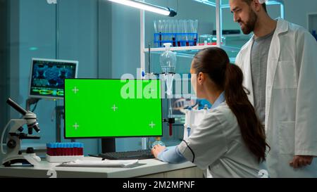 Female scientist working on computer with mock-up green screen in modern laboratory. Scientist doctor using various shelves with beakers, chemicals and different technical equipment is visible. Stock Photo
