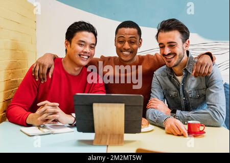 Group of three young male students and friends sitting in a cafè sitting at table looking at electronic device laughing. Stock Photo