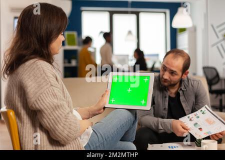 Back view of businesswoman sitting on couch using tablet with green screen talking with colleague while team working on background. Multiethnic coworkers planning project on chroma key display Stock Photo