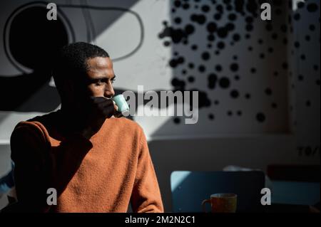 Side view young African handsome man sitting in a cafe drinking coffee with dramatic light. Stock Photo
