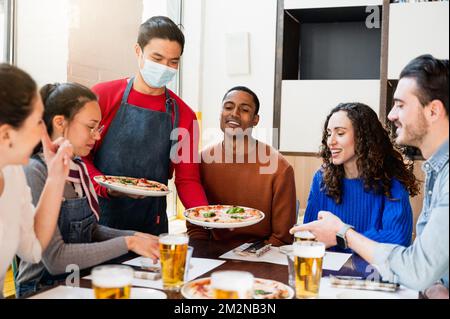Asian young waiter wearing protective face mask serving pizza to a multiracial group of young friends sitting at table restaurant. People with happy s Stock Photo