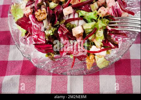 Fresh red and green salad leaf with sliced beetroot, tofu cube and walnut on glass plate, checkered tablecloth, closeup. Stock Photo