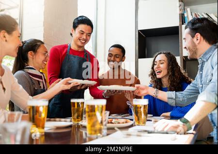 Multiracial happy group of young friends eating and drinking beer sitting at table in restaurant. Waiter serving pizza. Stock Photo