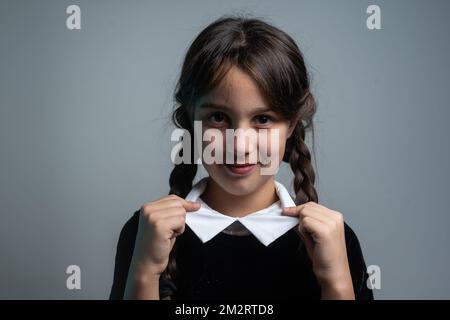 Portrait of little girl with Wednesday Addams costume during Halloween. Serious expression and dark atmosphere with dark background. Stock Photo