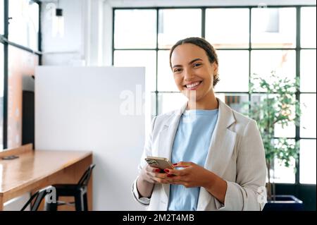 Attractive positive mixed race business lady, top manager of a company, standing in modern office, uses her smart phone, messaging in social media, answers email, looks at camera, smiling friendly