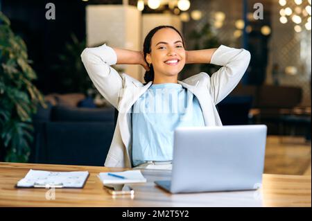Calm relaxed positive brazilian or hispanic young woman, secretary, manager, financial director, sitting at a desk in a modern office, resting during work time, looking at the camera, smiling friendly Stock Photo