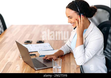Side view at positive, confident mixed race woman, with headset, works as call center agent, support service operator, helpdesk worker, consult clients online, makes recommendations, resolves issues Stock Photo