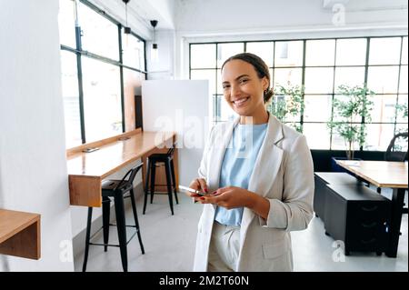 Online messaging. Positive latino business lady, manager of a company, standing in modern office, uses her mobile phone, messaging in social media, answers email, looks at camera, smiling friendly
