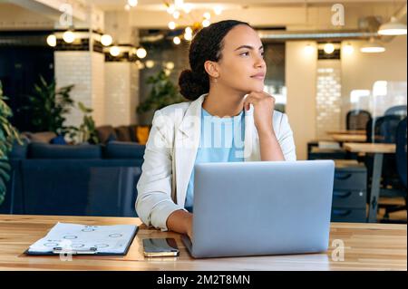 Calm beautiful clever elegant brazilian or hispanic young woman, secretary, manager, sitting at a desk in a modern office, looking away thoughtfully, thinking about strategy, dreaming about vacation Stock Photo
