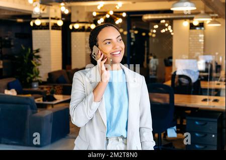 Successful business lady have smartphone conversation with a client, standing in the office, solving work issues, arranging a meeting, looking away, smiling friendly. Mobile phone conversation Stock Photo