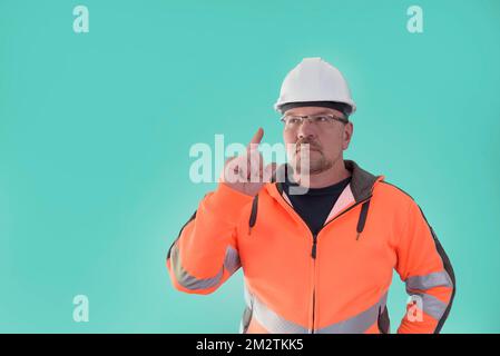 A male builder, thinking, finger up, in work clothes and a construction helmet, on a light green background. Stock Photo