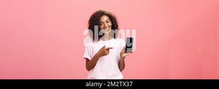 Suggest this model of phone. Happy charming african american curly-haired woman in stylish outfit holding smarpthone and pointing at device screen Stock Photo