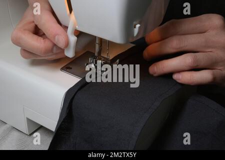 Seamstress sews clothes made of black cloth on a sewing machine. Work by the light of the built-in hardware lamp. Presser foot and steel needle with l Stock Photo