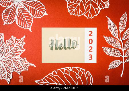 Hello 2023 text on note paper and paper leaves on red background. Stock Photo