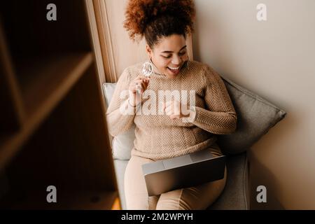 Young beautiful happy smiling african american woman adjusting her earrings while having a video conference while sitting on the windowsill Stock Photo