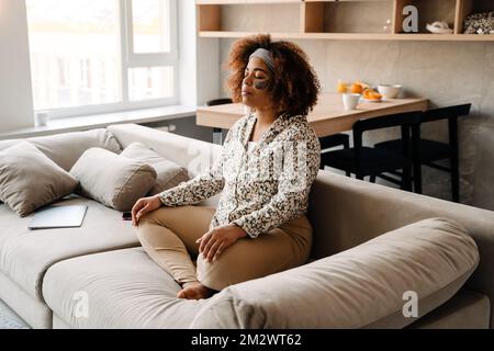 Young beautiful calm african woman with eye patches sitting on sofa in lotus pose and meditating Stock Photo
