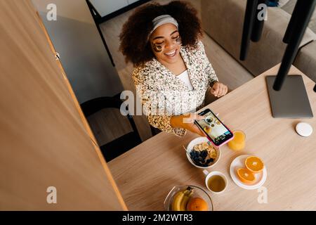 Top view of beautiful smiling african american woman taking photo of her breakfast, oatmeal with fruits, orange juice and tea Stock Photo
