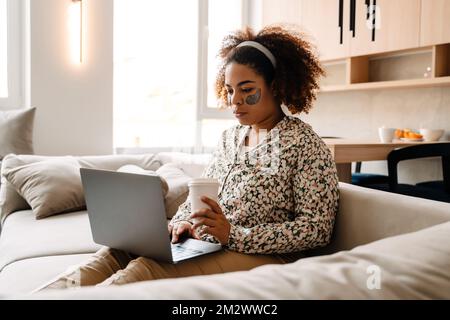 Young african american woman drinking coffee and using laptop while sitting on sofa at home Stock Photo