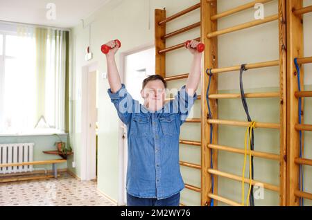 a man with down syndrome does sports with dumbbells in his hands. Stock Photo