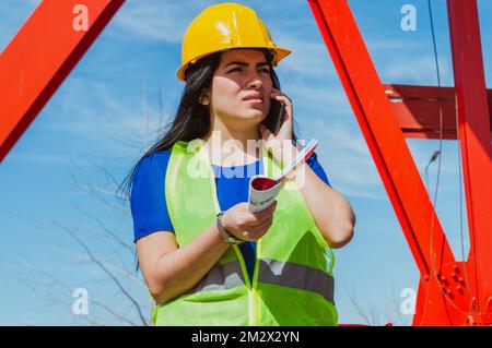 young latina caucasian female engineer outdoors, wearing yellow hardhat and safety vest, confused on a phone call talking to her boss, holding workboo Stock Photo