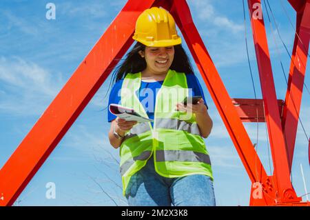 young caucasian latin woman engineer in a construction outdoors, she is smiling reading message and notifications on her phone, using mobile apps and Stock Photo