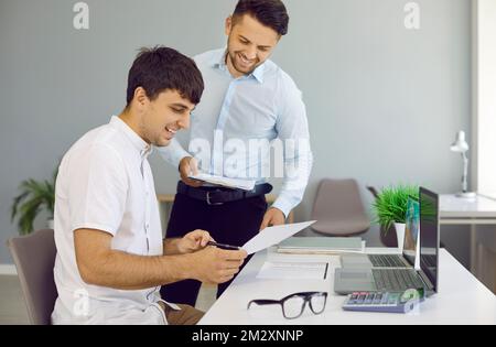 Cheerful colleagues are reviewing and discussing paper documents while working in office. Stock Photo