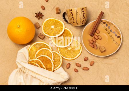 Coffee cup with saucer. Cinnamon stick and nuts. Slices of dried orange in fabric bag. Fresh orange. Flat lay. Beige paper background. Stock Photo