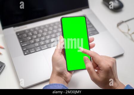 woman using smartphone with green screen over laptop in office. Stock Photo