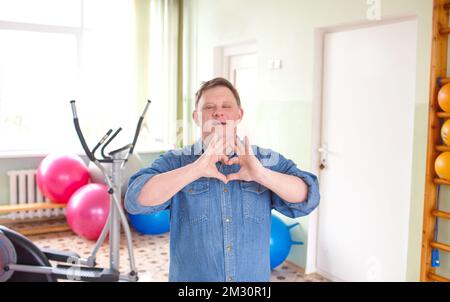 A young man with Down syndrome, dressed in a casual denim jacket smiling in the shape of a heart symbol with hands. romantic concept Stock Photo