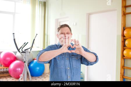 A young man with Down syndrome, dressed in a casual denim jacket smiling in the shape of a heart symbol with hands. romantic concept Stock Photo