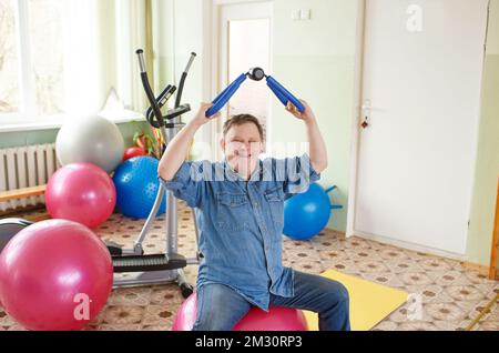 a man with down syndrome sits on a large inflatable ball with a gymnastic stick in a rehabilitation center Stock Photo