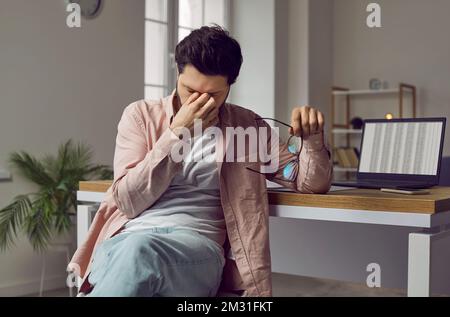 Tired, stressed man with eye strain sitting at working desk with laptop computer Stock Photo