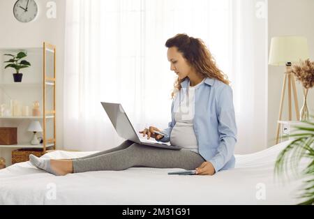 Focused young pregnant woman on maternity leave working online at home using laptop. Stock Photo