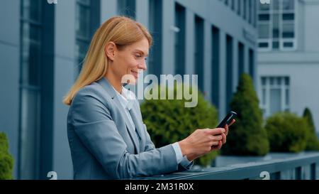 Smiling attractive woman standing outdoors holding phone checking email making order in online store happy businesswoman communicating in social Stock Photo