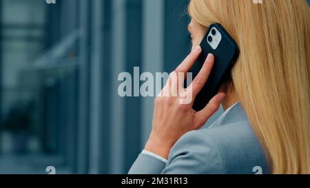 Back view close-up blonde caucasian businesswoman talking on mobile phone answering business call remotely communicates with client using smartphone Stock Photo
