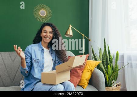 Happy woman buyer at home satisfied with delivery of goods from online store, Hispanic woman sitting on sofa and looking at camera holding box with goods in hands. Stock Photo