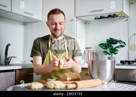 Attractive caucasian bearded chef baker in green apron working with dough on wooden cutting board with rolling pin on foreground. Working at home kitchen concept, homemade baking. High quality photo Stock Photo
