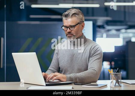 Portrait of a senior gray-haired man, a businessman, director, founder, who is concentrating on working in the office at a desk with a laptop. Stock Photo