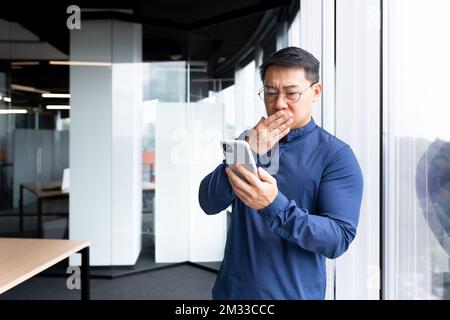 Upset asian man in office at work reading bad news from phone online, businessman in shirt near window working inside office, using smartphone. Stock Photo