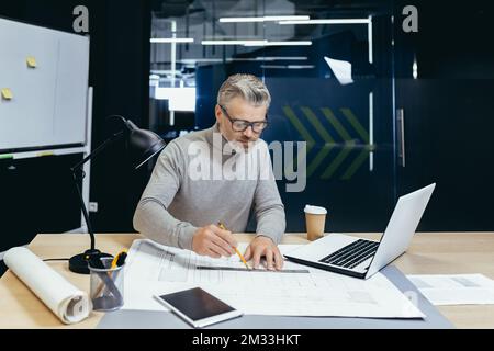 General photo of the workplace of a gray-haired senior man, a designer, architect, engineer. He sits at the desk in the office. He draws a project, a plan with a pencil and a ruler on a roll of paper. Stock Photo