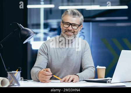 Portrait of an older gray-haired male teacher. He sits at a desk with a laptop, looks at the camera, smiles, holds a pencil in his hands. Teaches online, remotely. Stock Photo