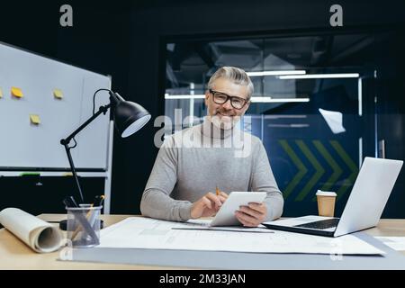 A senior gray-haired man, architect, designer, engineer. Sitting in the office with a laptop and paperwork. He holds a notebook with a pencil in his hands. He looks at the camera and smiles. Stock Photo