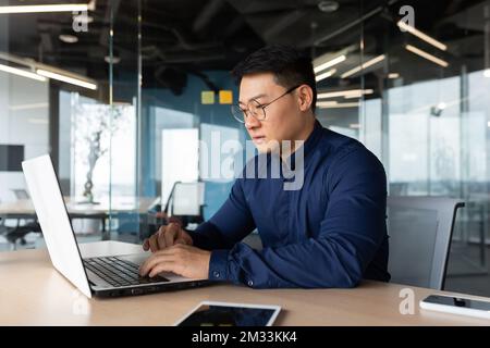 Serious and thoughtful businessman working inside office sitting at table using laptop at work, mature asian boss in shirt thinking and typing on keyboard. Stock Photo