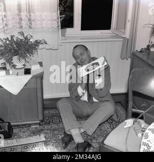 1960s, historical, a man in a suit & tie sitting on the floor of a front room of a house holding a carton of king size cigarettes, acting like he is heaven! Stock Photo