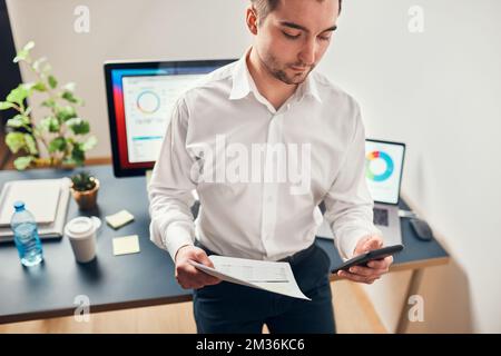Man entrepreneur having business  conversation on mobile phone. Businessman working in office holding documents and smartphone. Man standing at desk w Stock Photo