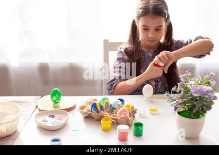 Colorful painted eggs in a tray stand on the table, the girl opens a jar of paint in the background. Stock Photo