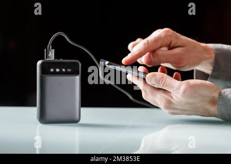 Black powerbank with a black cord charging  smartphone in a male hands on a black. Extra power supply. Minimalist image Stock Photo